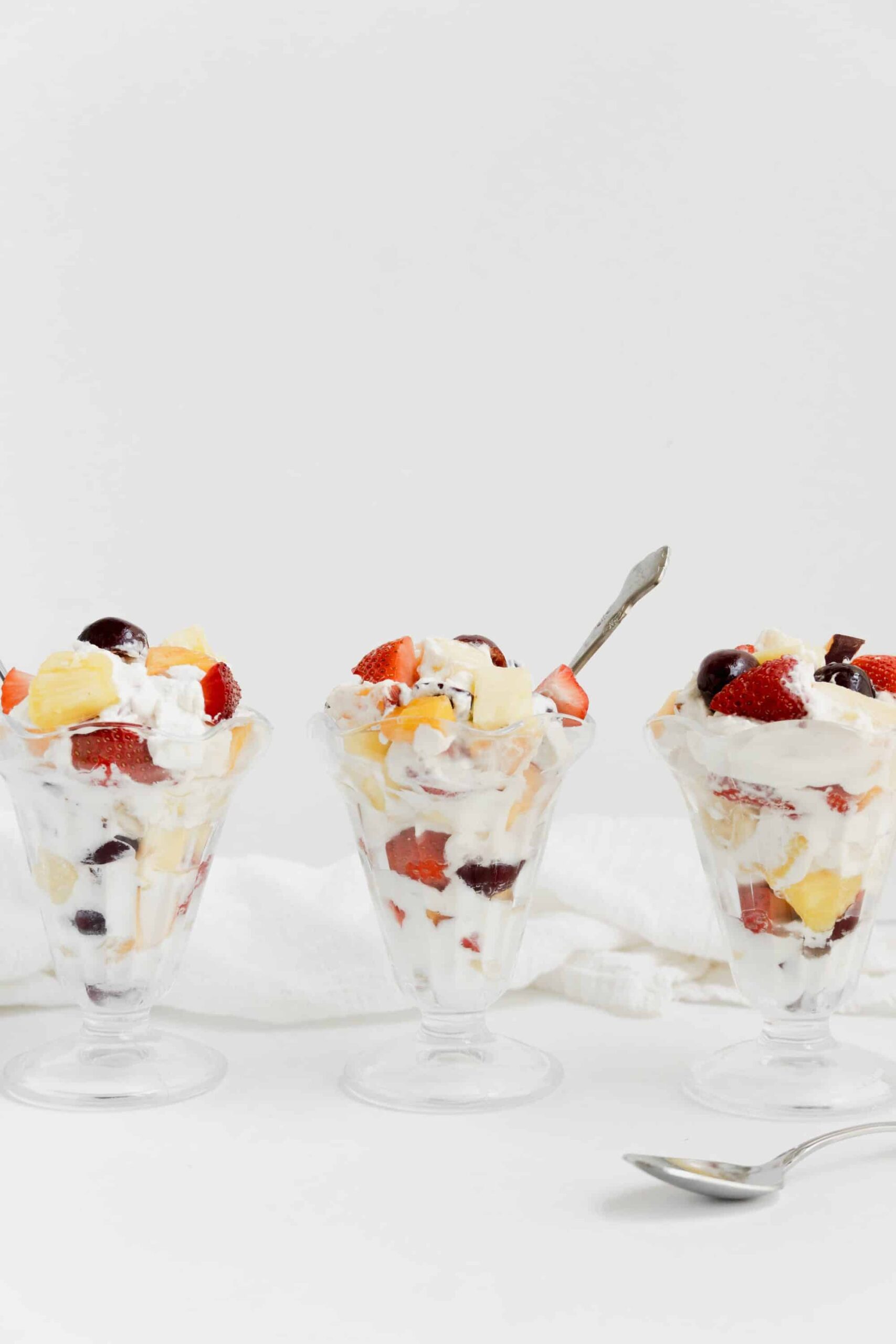 Three sundae glasses of whipped cream fruit salad with a white backrdop.
