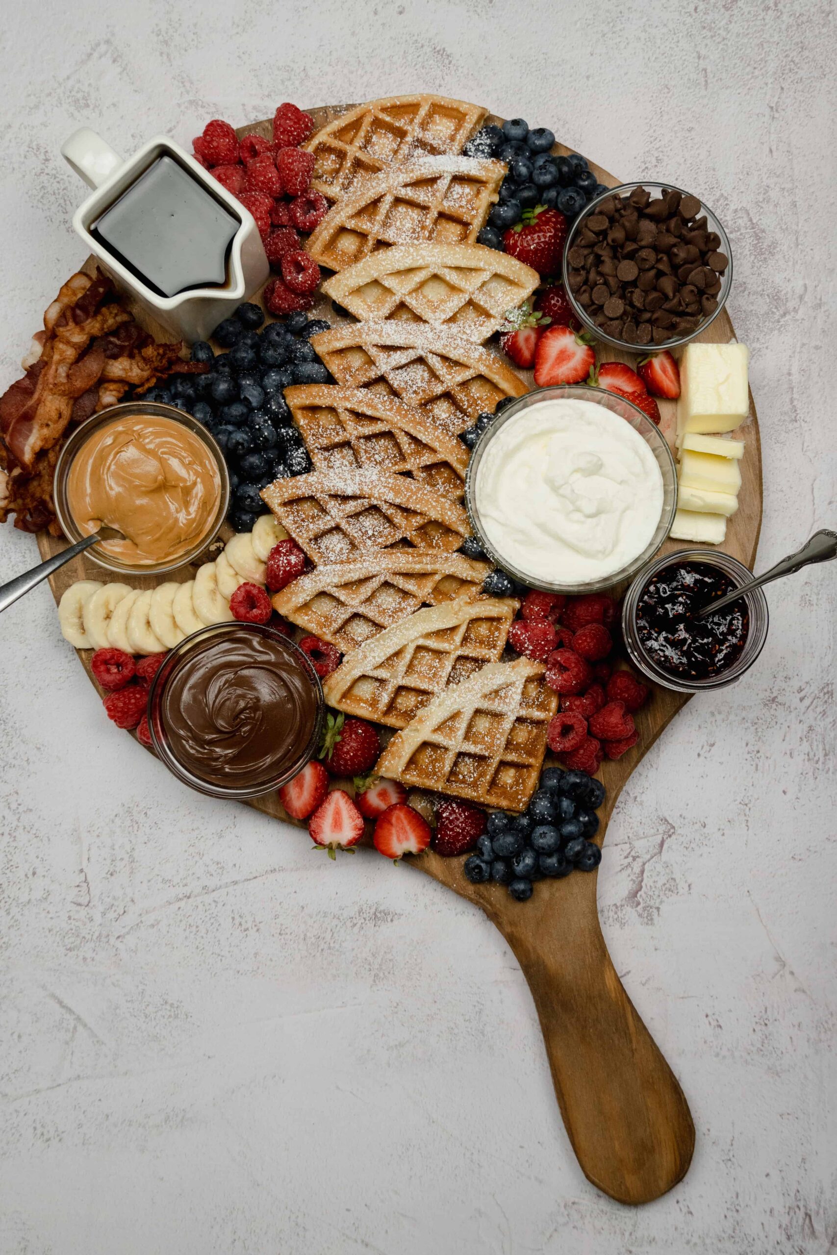Waffles, fruit, syrup, jam, chocolate, bacon, and butter on a charcuterie board with powdered sugar on top.